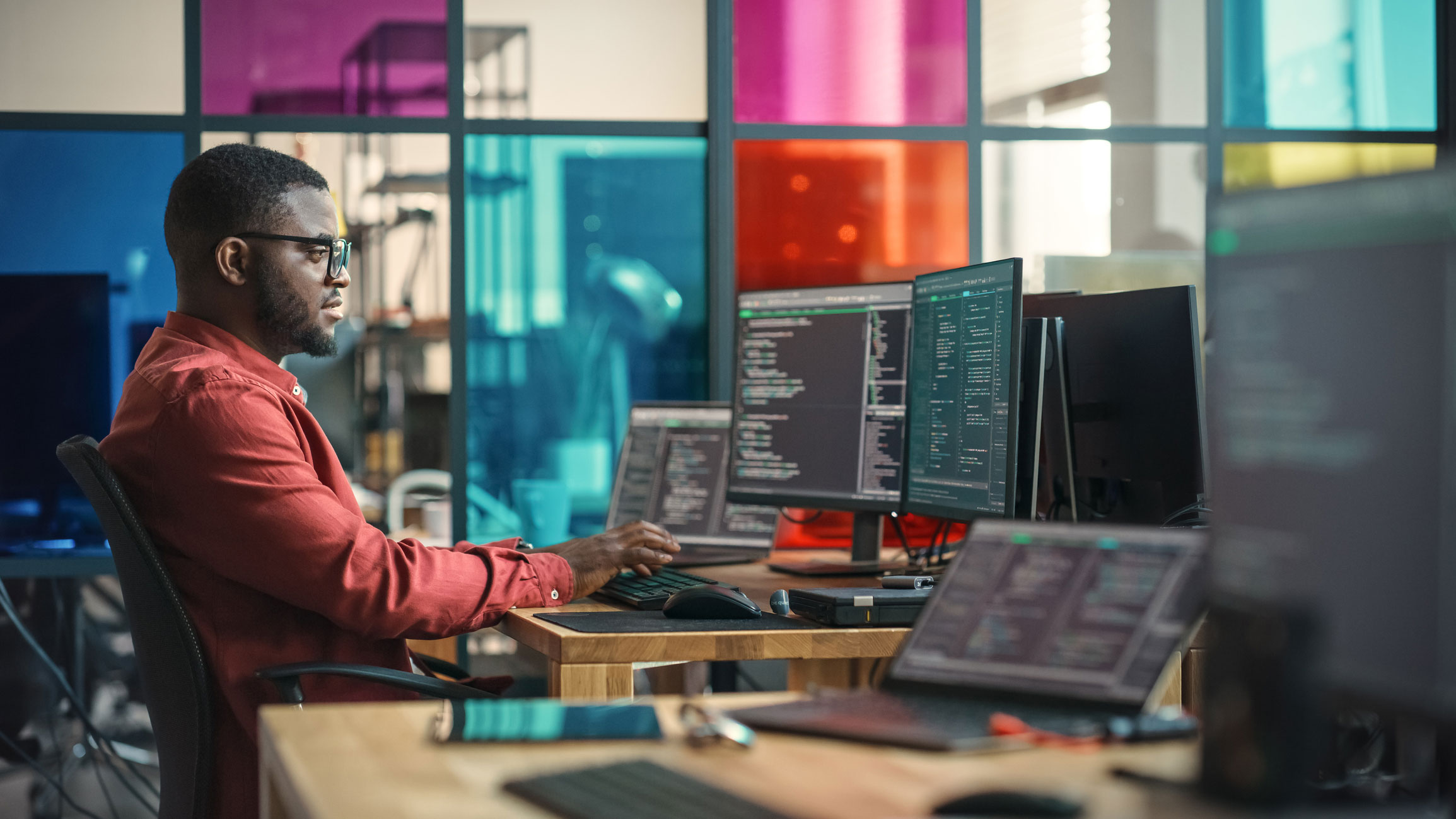 An IT Engineer sits at a computer desk observing code on multiple screens.