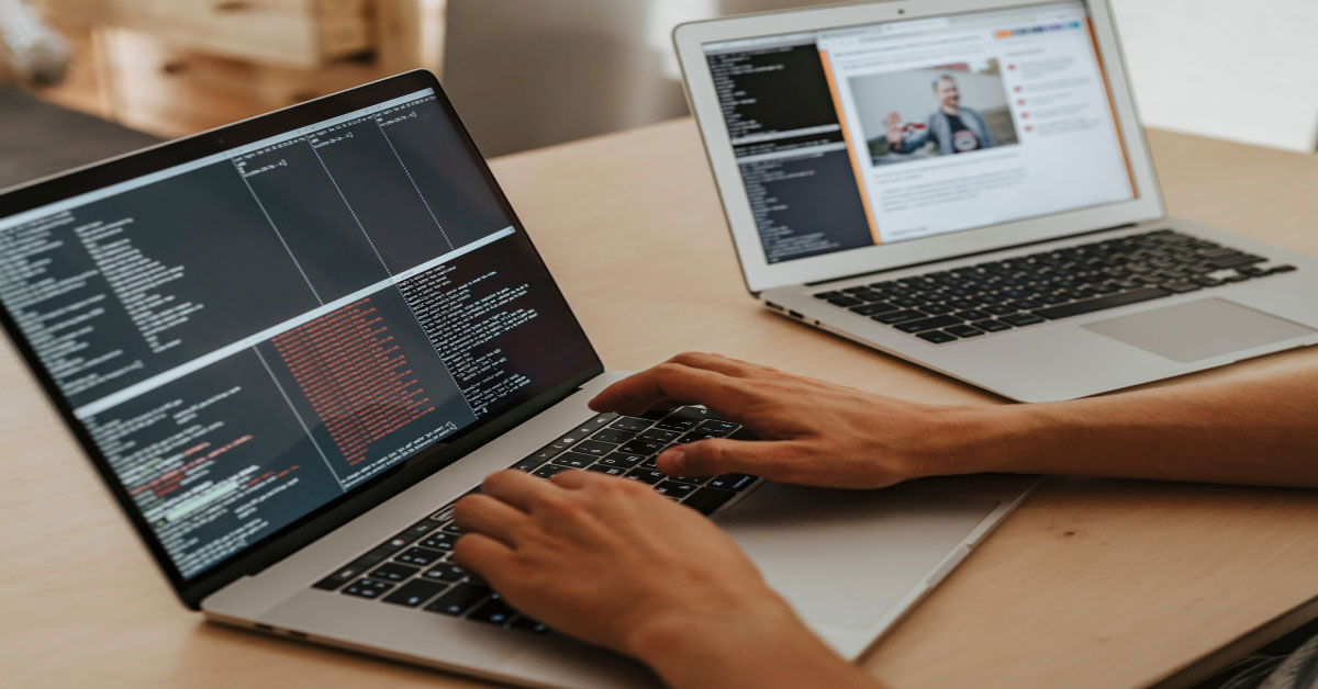 A man is performing web services testing by typing testing code into one silver laptop while running the test code on the website of another silver laptop.