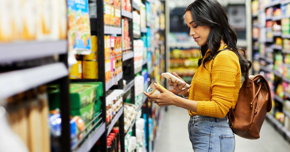 Woman stands in a grocery aisle with a canned product in her hand while using her phone to check the price of the item.